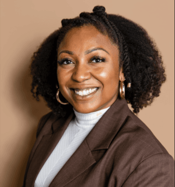 Smiling Black woman with curly hair, gold hoop earrings, white turtleneck, and brown blazer.