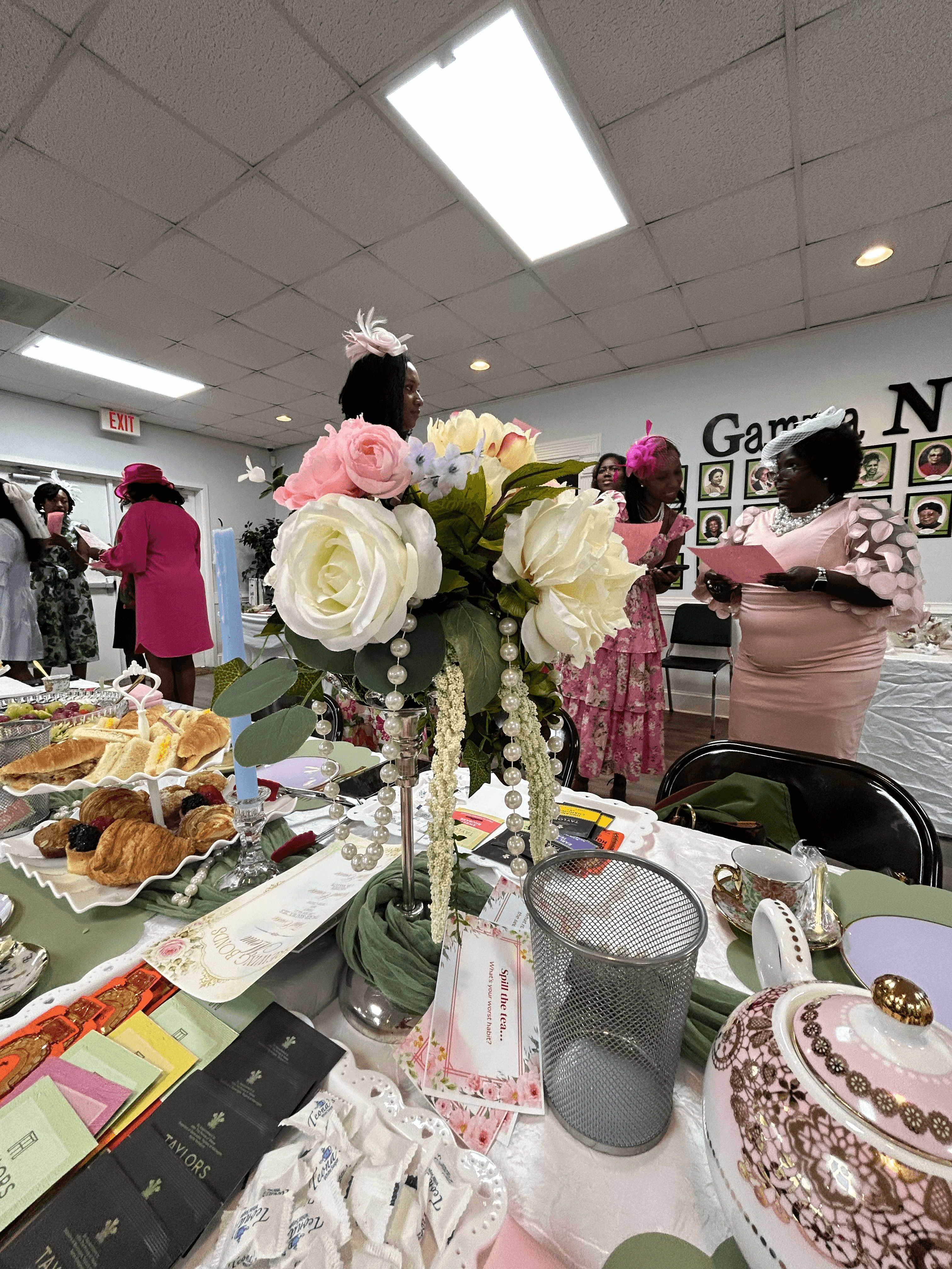 Women in formal dresses and hats attend a tea party featuring floral centerpieces and pastries.